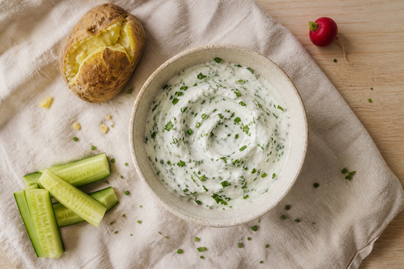Cremiger Kräuterquark mit Schnittlauch und Petersilie neben Gurkensticks, Ofenkartoffel und einem Radieschen